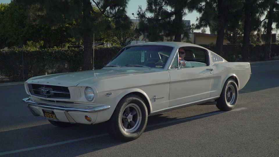 White 1960s Ford Mustang coupe with a man driving.