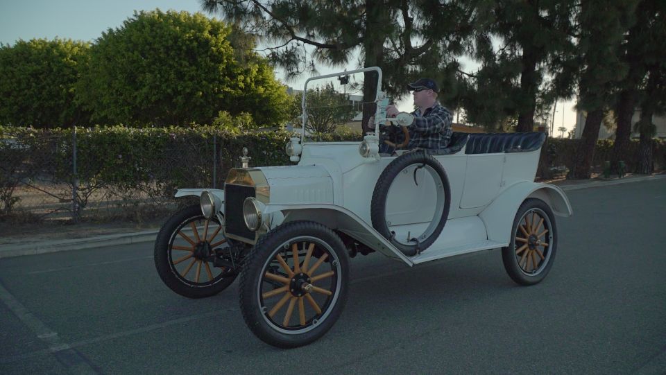 White vintage touring car with wooden spoked wheels and black convertible top, driven by a man.