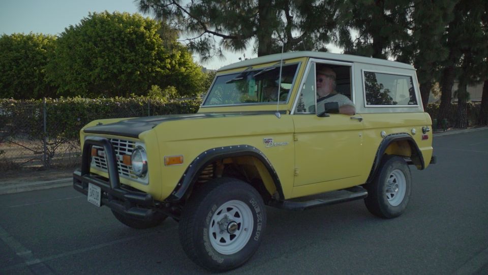 Yellow 1970s Ford Bronco with a man driving, parked outdoors.