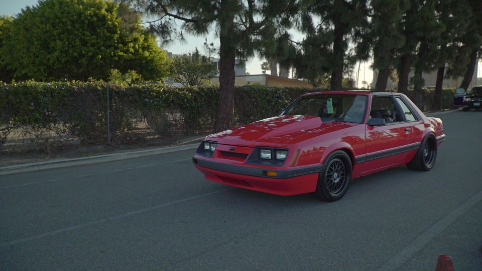 Red 1987-1993 Ford Mustang hatchback with hood scoop and black wheels parked curbside.
