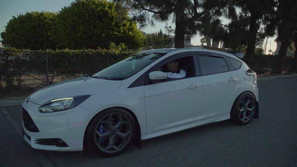White Ford Focus ST with aftermarket wheels and spoiler, driver visible inside.
