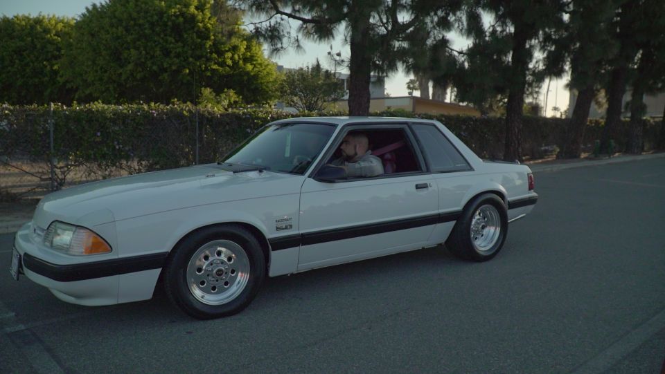 White 1990s Ford Mustang coupe with aftermarket wheels and a pink racing seatbelt, parked on a road.