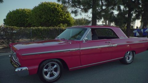 1993 Ford Taurus - A pink 1965 Ford Galaxie 500 coupe is parked on a street with trees and a fence in the background.