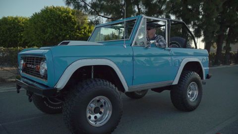 1993 Ford Taurus - A man drives a lifted blue first-generation Ford Bronco on a paved road with trees in the background.