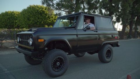 1993 Ford Taurus - A modified black Ford Bronco from the early 1970s is driven on a paved road with trees in the background.