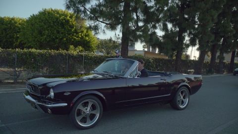 1993 Ford Taurus - A dark purple 1965 Ford Mustang convertible is seen driving down a street with trees and a hedge in the background.