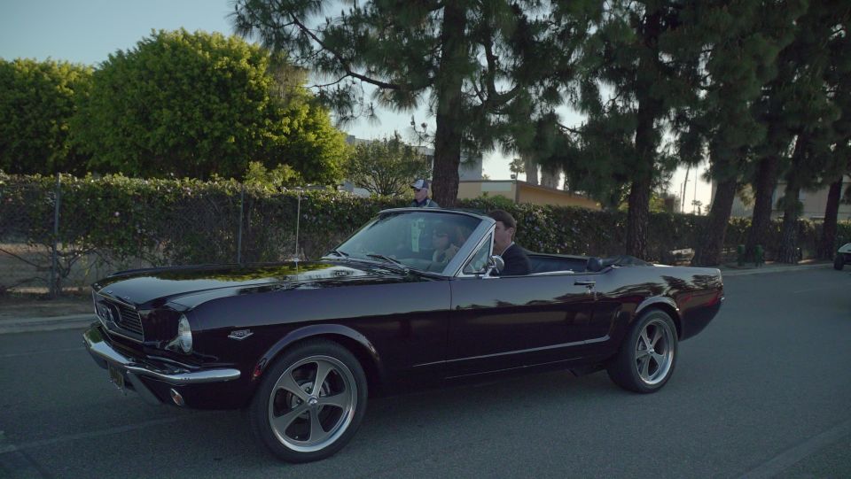 Dark purple 1965 Ford Mustang convertible, driver and passenger visible, driving on a street