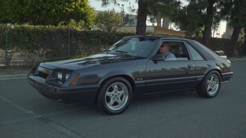 1993 Ford Taurus - A dark gray 1987 Ford Mustang GT coupe is driven down a street, with trees and a hedge in the background.