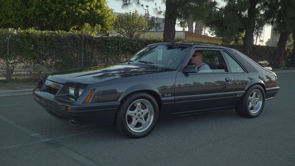 Dark gray 1987 Ford Mustang GT coupe with a driver, seen from the front 3/4 angle
