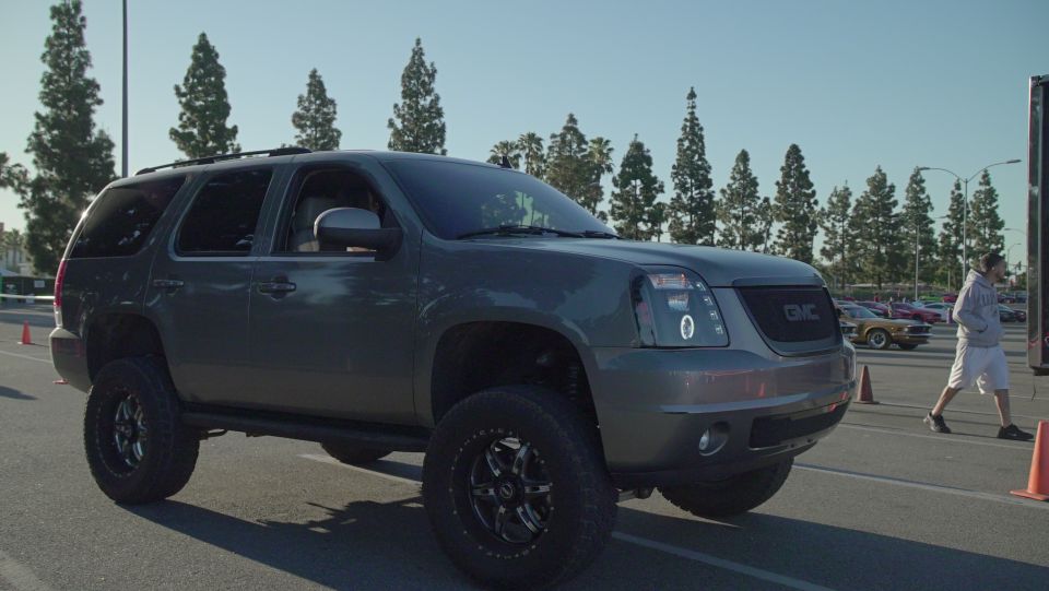 Lifted gray GMC Yukon XL with custom headlights and grille. Other cars and a person walking in the background