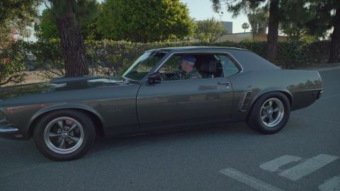 1993 Ford Taurus - A driver in a blue hat sits behind the wheel of a dark gray 1969 Ford Mustang Mach 1.