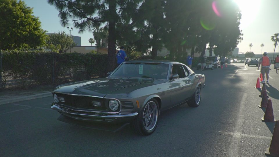 Gray 1970 Ford Mustang driving on a street with trees and people in the background