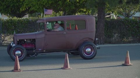 1993 Ford Taurus - A man drives a vintage hot rod sedan, marked by traffic cones, with an American flag flying from its antenna.