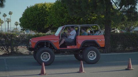 1993 Ford Taurus - A lifted orange Ford Bronco with a roll cage drives past traffic cones on a sunny day.