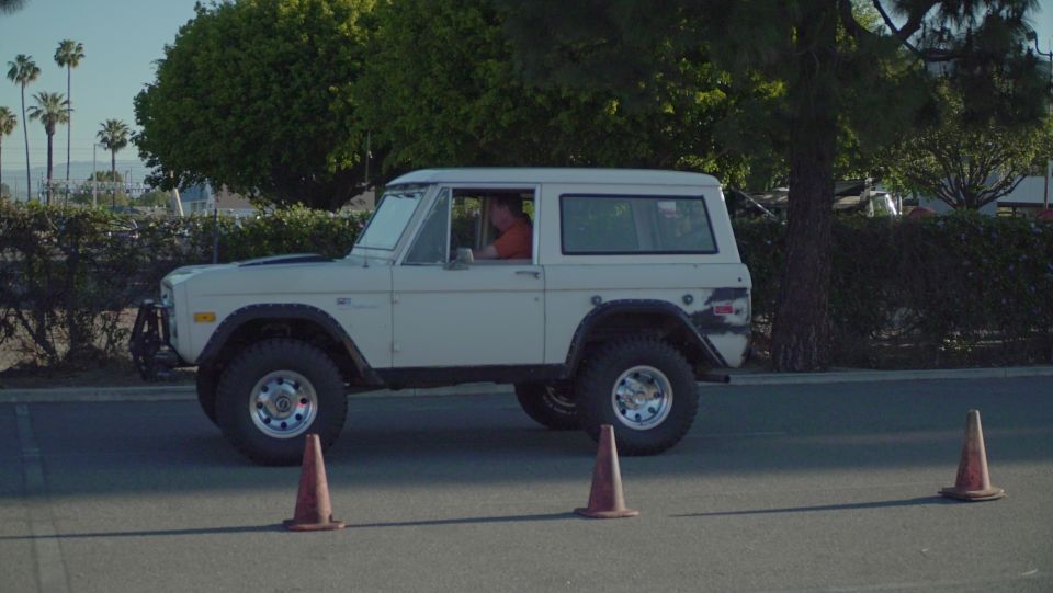 White 1978 Ford Bronco driving past orange cones on asphalt