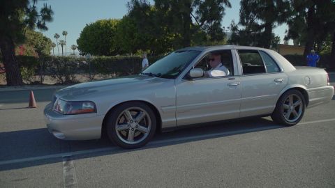 1993 Ford Taurus - A silver sedan is parked on an asphalt lot, with a driver visible inside. Trees and a traffic cone are in the background.