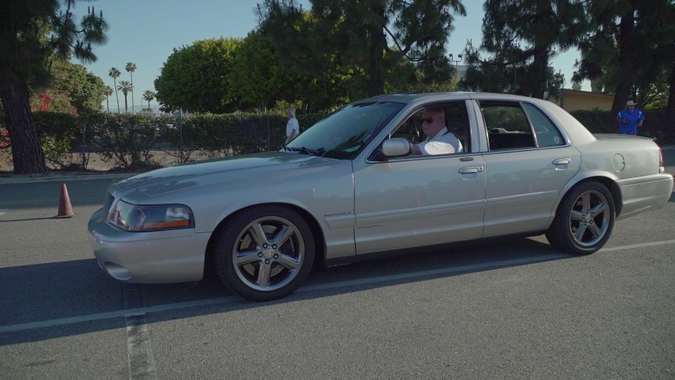 Silver sedan with a man driving, parked on asphalt with trees and a cone nearby