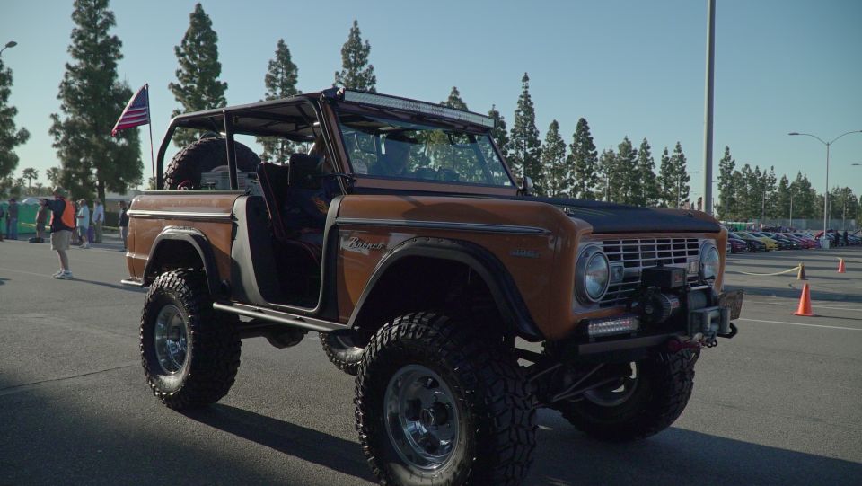 Lifted orange first-gen Ford Bronco with a roll cage and spare tire in the back