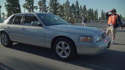 1993 Ford Taurus - A man in an orange vest looks out over a silver sedan parked in a lot with trees in the background.