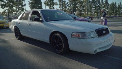 1993 Ford Taurus - A white sedan with aftermarket black wheels is parked in a lot, with other vehicles and people in the background.