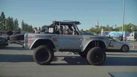 1993 Ford Taurus - A modified silver Ford Bronco with racing decals is parked next to a modern silver car at an outdoor event.