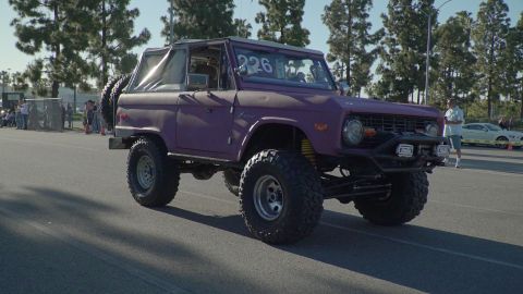 1993 Ford Taurus - A modified purple Ford Bronco is driven on a paved surface, with spectators and other vehicles in the background.