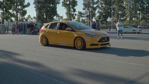 1993 Ford Taurus - A yellow hatchback car drives on a paved surface with trees and people in the background.