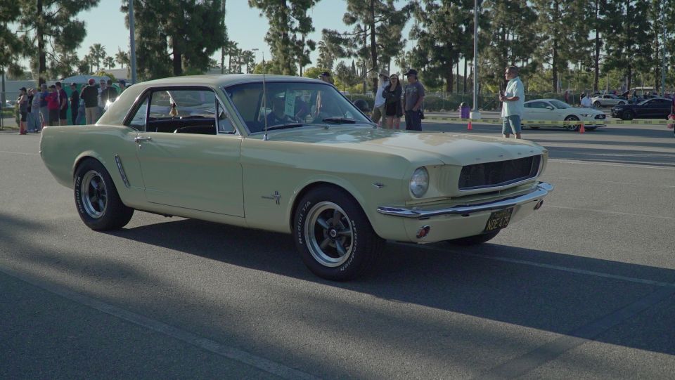 Pale yellow 1965 Ford Mustang coupe, 3/4 front view. Other cars visible in background