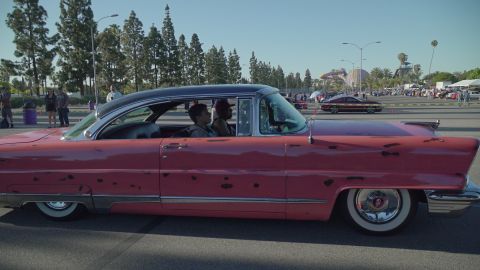 1993 Ford Taurus - A classic pink car with a two-tone roof is seen from the side, with people inside and other vehicles in the background.