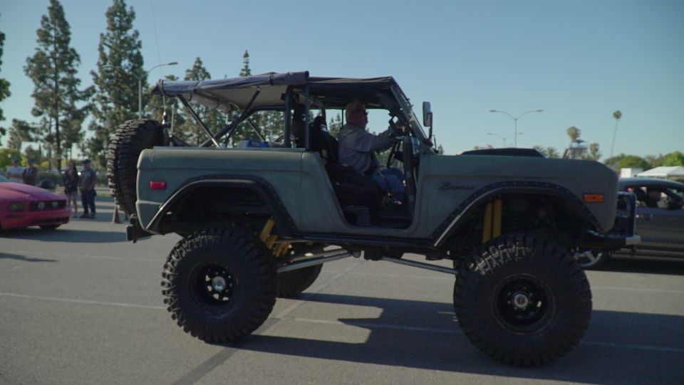 Lifted olive green Ford Bronco with roll cage and off-road tires, driver visible.