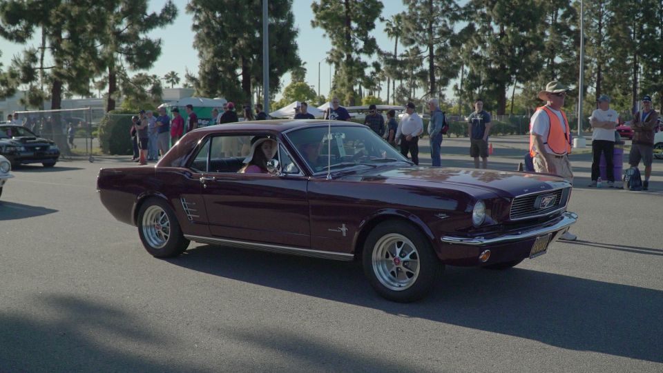Maroon 1960s Ford Mustang coupe with a woman in the driver's seat, parked outdoors.