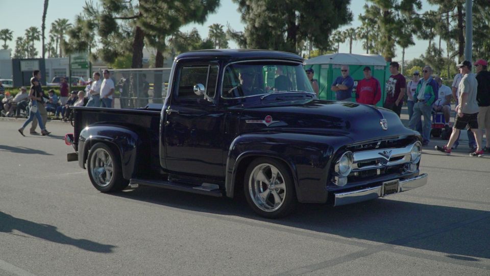 Dark blue classic Ford pickup truck driving on a road, with people watching.