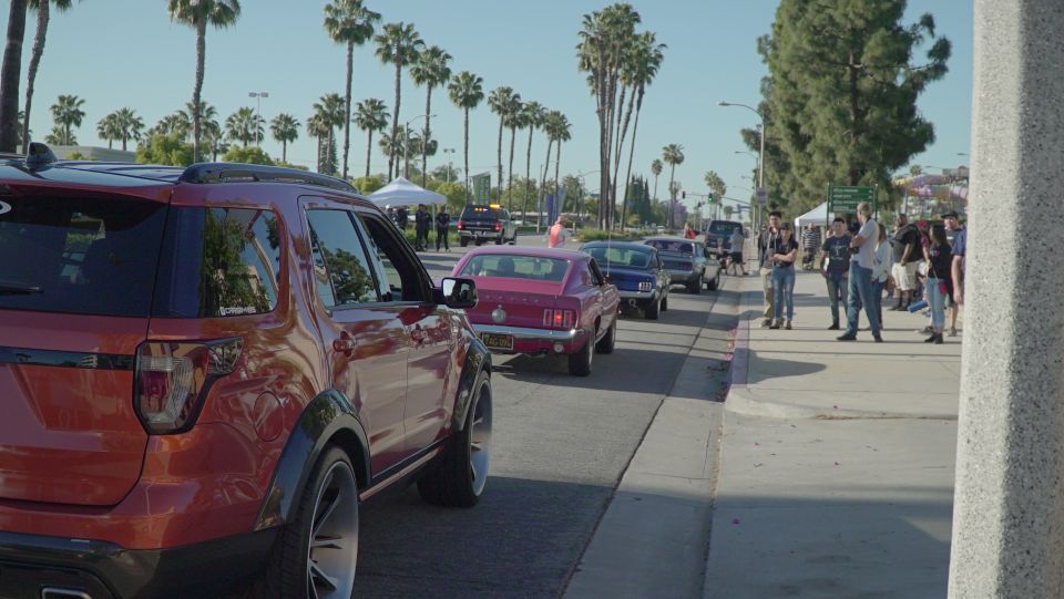 Classic cars in a parade with palm trees and people watching. Red SUV in foreground.