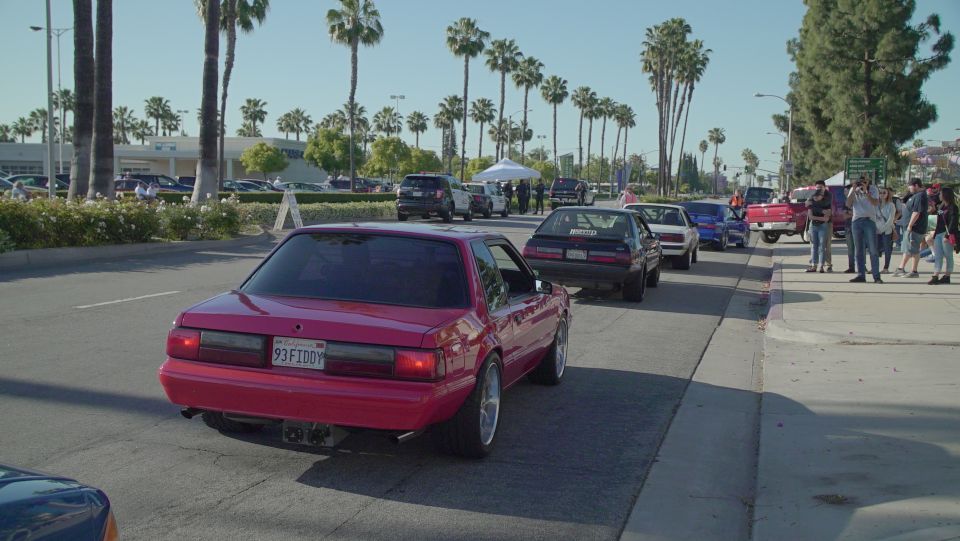 Pink 1993 Ford Mustang coupe from rear three-quarter view, driving on a street with palm trees.
