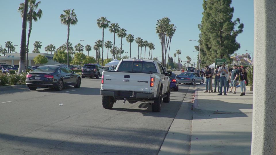 White Ford F-150 Raptor pickup truck driving down a street with palm trees and people on the sidewalk.