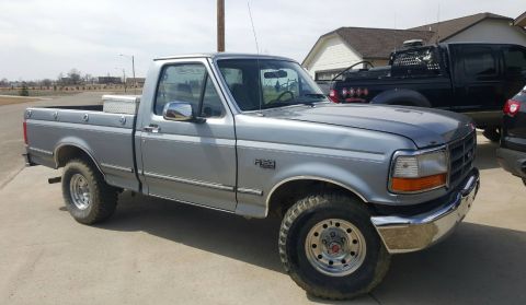 1995 Ford F-150 - A silver 1990s Ford F-150 pickup truck is parked on a concrete surface in front of a house. The truck has a single cab, a long bed, and chr…
