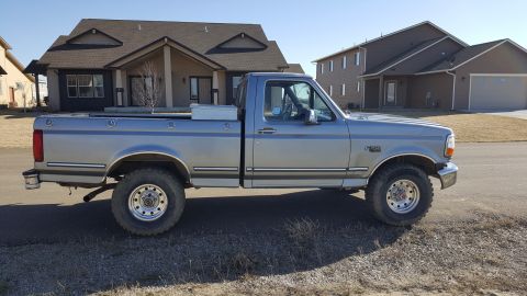 1995 Ford F-150 - A silver 1990s Ford F-150 pickup truck is parked on a residential street in front of two houses. The truck has a standard cab, a long bed,…