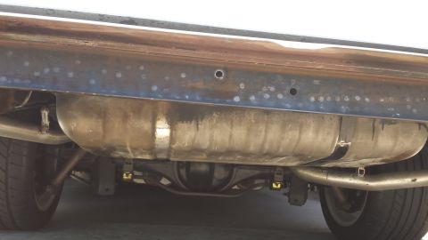 1976 Ford Ranchero - The underside of a vehicle shows a large fuel tank mounted above the rear differential and exhaust system. The vehicle's frame has numerous…