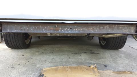 1976 Ford Ranchero - The underside of a vehicle showing the rear bumper support bar, exhaust system, and rear tires. A piece of cardboard is visible in the fore…