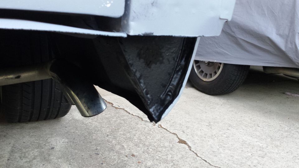 Close-up of a car's rear bumper area, showing an exhaust pipe and a tire. Another vehicle covered by a tarp is visible