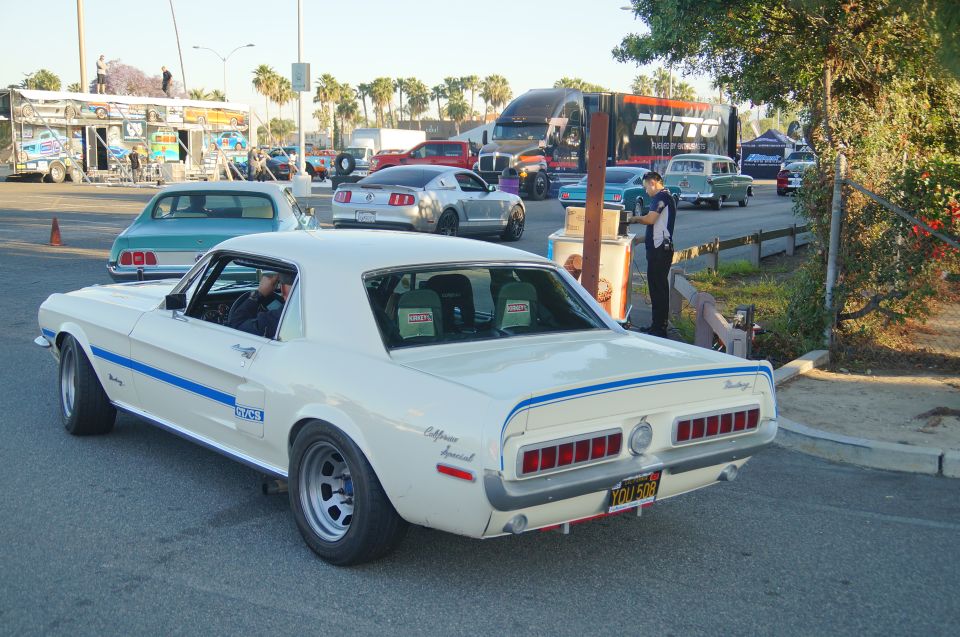 White 1960s Ford Mustang coupe with blue stripe, parked outdoors with trucks and other cars.
