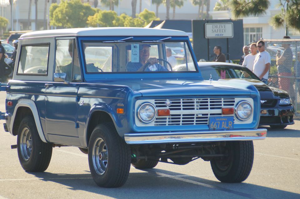 Blue first-generation Ford Bronco with white roof, driver visible.