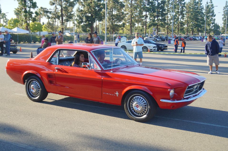 Red classic Ford Mustang coupe with two people inside, parked at an outdoor event.