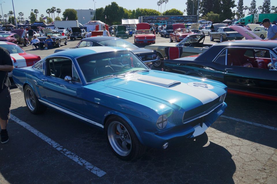 Blue 1960s Ford Mustang coupe with white racing stripes, parked at a car show.
