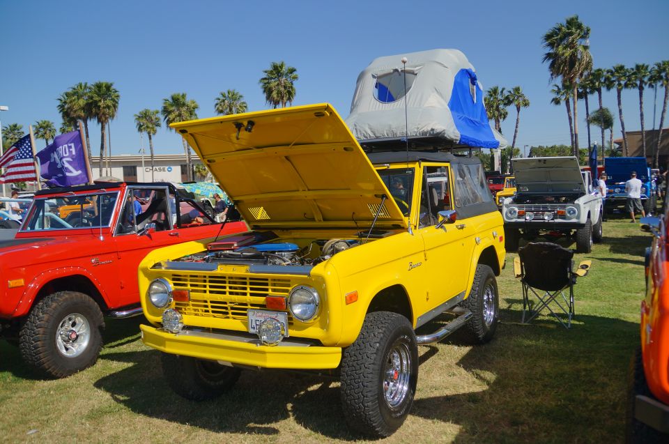 Yellow first-generation Ford Bronco with open hood and rooftop tent parked on grass.