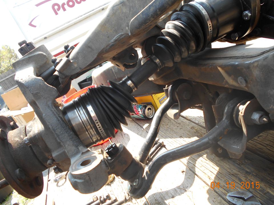 Close-up of vehicle front suspension with CV axle, steering knuckle, and control arms.