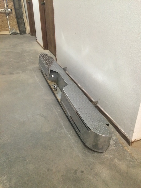 1981 Ford Bronco - A chrome aftermarket rear bumper with diamond plate step is leaning against a white wall on a concrete floor. The bumper has a visible trai…