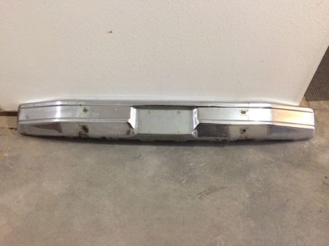 1981 Ford Bronco - A chrome rear bumper is shown mounted to a white wall, resting on a concrete floor. The bumper has a rectangular cutout in the center for a…