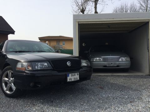 1999 Ford Fairlane - A black sedan is parked in front of a garage where a silver sedan is parked inside. The black sedan has a license plate that reads "BÜR MV…