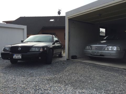 1999 Ford Fairlane - A black Mercury Grand Marquis is parked in front of a garage, with a silver Ford Fairlane visible inside the garage. The Mercury has a Germ…